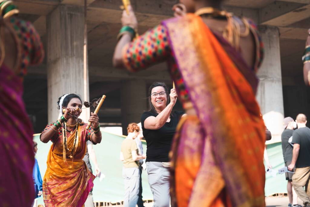 A group of dancers in vibrant, colorful traditional attire performing in front of a cruise ship. They are engaging with onlookers, including a smiling woman in casual clothes who appears to be participating in the dance.