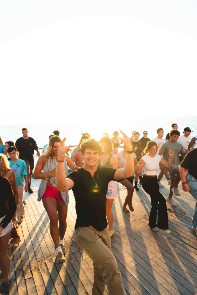 A crowd of people are gathered in rows on the deck of a ship, learning a dance. Each has one leg raised, bent at the knee; a young man at the front of the crowd raises his arms and smiles at the camera. They are all lit from behind by a setting sun.