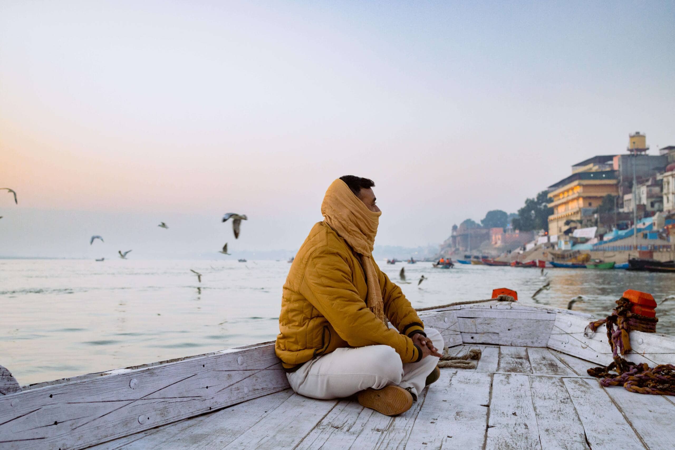 A young man in a yellow jacket sits in a wooden boat on a river, looking toward the buildings along the shore. There is a yellow pashmina scarf wrapped around the lower half of the man's face. Seagulls fly in the background.