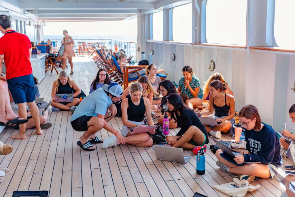 Groups of students sit together on the wooden deck of a ship, huddled in discussion or working independently on laptops. The sparkling ocean is just visible in the background behind them.
