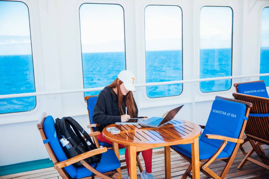 A woman with long, red hair sits at a round wooden table surrounded by blue-cushioned folding chairs, writing on an iPad with a stylus while a laptop sits open in front of her. A blue ocean is visible through porthole windows behind her.